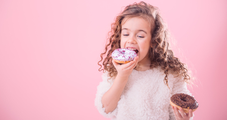 Portrait of a little joyful cute curly girl who eats donuts, on a pink background, a place for textの写真素材