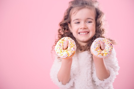 Portrait of a little smiling girl with curly hair and appetizing donuts in her hands, on a pink background, a place for textの写真素材