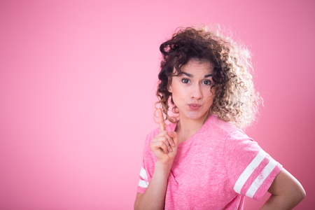 Portrait of young surprised girl with curly hair in loose pink t-shirt shows finger to the side isolated on pink background, place for text, emotion conceptの写真素材