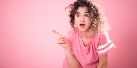Portrait of young surprised girl with curly hair in loose pink t-shirt shows finger to the side isolated on pink background, place for text, emotion conceptの写真素材
