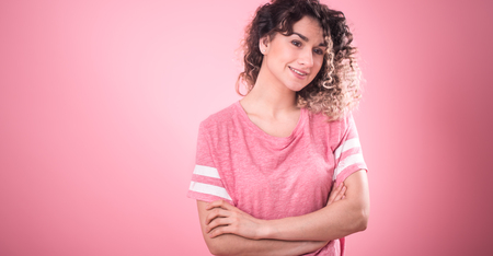 Portrait of young beautiful cheerful girl with curly hair and loose pink t-shirt isolated on pink background, place for text, woman concept and emotionの写真素材