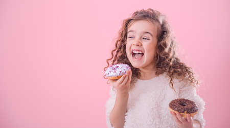 Portrait of a little joyful cute curly girl who eats donuts, on a pink background, a place for textの写真素材