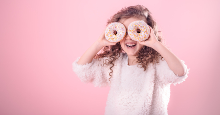 Portrait of a little smiling girl with curly hair and two appetizing donuts in her hands, closes her eyes with donuts, on a pink background, a place for textの写真素材