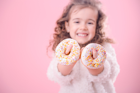 Portrait of a little smiling girl with curly hair and appetizing donuts in her hands, on a pink background, a place for textの写真素材