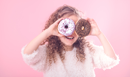Portrait of a little smiling girl with curly hair and two appetizing donuts in her hands, closes her eyes with donuts, on a pink background, a place for textの写真素材