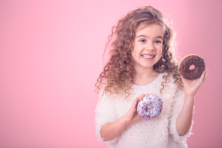 Portrait of a little smiling girl with curly hair and appetizing donuts in her hands, on a pink background, a place for textの写真素材