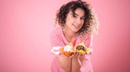Portrait of a cheerful young curly-haired girl with cupcakes in her hands, in a loose pink t-shirt isolated on a pink background, a place for text, the concept of emotions and healthy eatingの写真素材