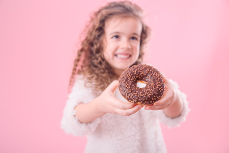 Portrait of a little smiling girl with curly hair and appetizing donuts in her hands, on a pink background, a place for textの写真素材