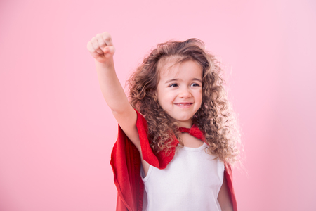 The concept of children, a smiling curly-haired girl playing a super hero, pointing up , isolated on pink backgroundの写真素材