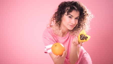 Portrait of a young curly-haired girl when choosing the right food with orange and cupcake in hand, in a loose pink t-shirt isolated on a pink background, a place for text, the concept of emotions and healthy eatingの写真素材
