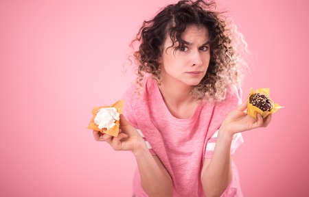 Portrait of a young curly-haired girl with cupcakes in her hands, in a loose pink t-shirt isolated on a pink background, a place for text, the concept of emotions and healthy eatingの写真素材