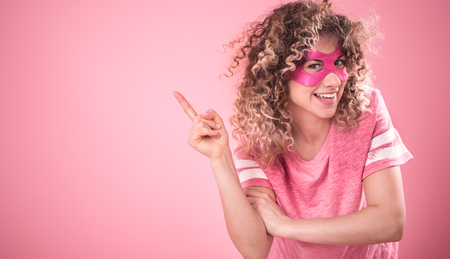 Women concept, Portrait of beautiful curly-haired young girl in superhero mask shows finger to the side, in free pink t-shirt on isolated pink background, place for textの写真素材