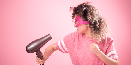 Hair care woman with Hairdryer, a curly-haired girl in the mask of a superhero with a Hairdryer in hand, in a free pink t-shirt isolated on pink background ,place for text , Concept of beauty and careの写真素材