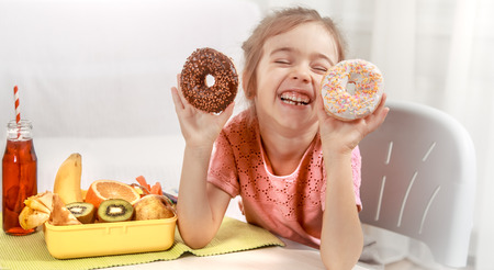Little beautiful cheerful girl eating a donut and lunch box with fruits at home at the table, the concept of children's foodの写真素材