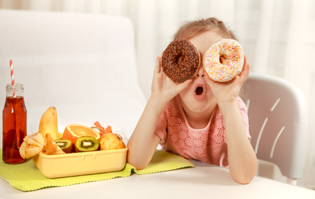 Little beautiful cheerful girl eating a donut and lunch box with fruits at home at the table, the concept of children's foodの写真素材