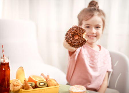 Little beautiful cheerful girl eating a donut and lunch box with fruits at home at the table, the concept of children's foodの写真素材