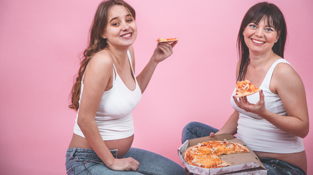 food concept, two beautiful pregnant women eating pizza on a pink background, in jeans and a white t-shirt, female friendshipの写真素材