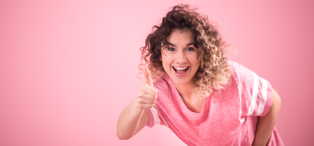 Portrait of young beautiful cheerful girl with curly hair and loose pink t-shirt isolated on pink background, place for text, woman concept and emotionの写真素材