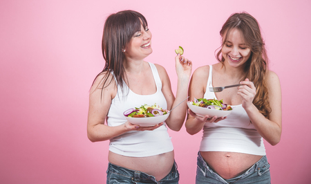 maternity concept, two pregnant women eating fresh salad on a pink colored background, in jeans and a white T-shirt, female friendshipの写真素材