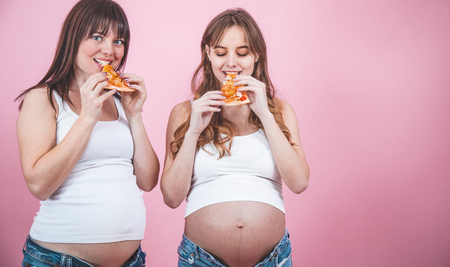 food concept, two beautiful pregnant women eating pizza on a pink background, in jeans and a white t-shirt, female friendshipの写真素材