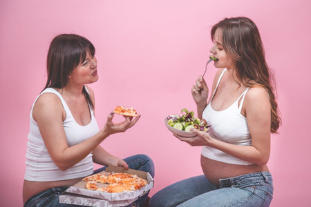 food concept, two beautiful pregnant women eating pizza on a pink background, in jeans and a white...