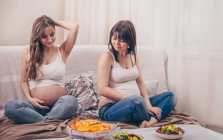 Maternity concept, two pregnant women eating pizza and salad at home on the sofa, friendship and healthy eatingの写真素材