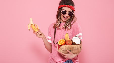 Horizontal portrait of hipster girl in stylish clothes with sunglasses, with healthy food, fruits, on pink background, lifestyle conceptの写真素材