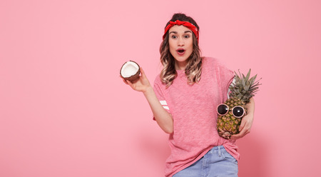Horizontal portrait of hipster girl in stylish clothes, with pineapple and coconut in her hands, on a pink background, lifestyle conceptの写真素材