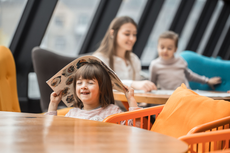 A charming little girl was sitting at a wooden table with a menu on her head in the restaurant. Leisure and communication in the restaurant.の写真素材