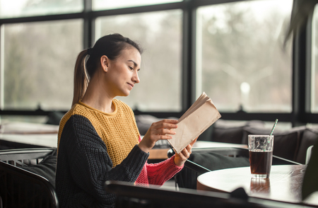 Young beautiful woman in orange sweater reading interesting book in cafe. The concept of readingの写真素材