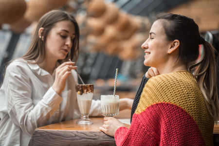 Two happy women are sitting in a cafe, drinking milkshakes, telling each other funny stories, being in a good mood, laughing happily. Best friends have fun together in the cafeの写真素材
