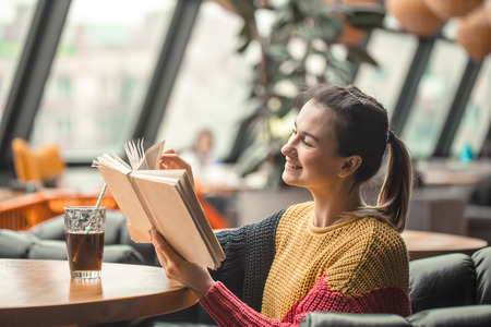 Young beautiful woman in orange sweater reading interesting book in cafe. The concept of readingの写真素材