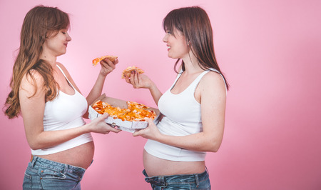 food concept, two beautiful pregnant women eating pizza on a pink background, in jeans and a white t-shirt, female friendshipの写真素材