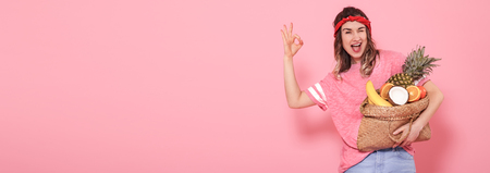 Beautiful young girl in pink t-shirt, holds a full bag of fruit straw on pink background. Shows hand OK. Place for text. Advertising conceptの写真素材