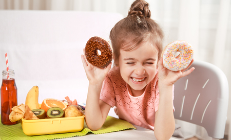 Little beautiful cheerful girl eating a donut and lunch box with fruits at home at the table, the concept of children's foodの写真素材