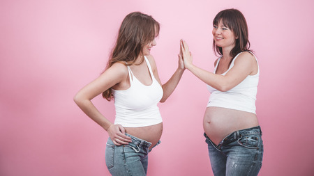 maternity concept, two beautiful pregnant women with an open tummy in jindes and white t-shirts on a colored background, female friendshipの写真素材