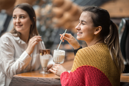 Two happy women are sitting in a cafe, drinking milkshakes, telling each other funny stories, being in a good mood, laughing happily. Best friends have fun together in the cafeの写真素材
