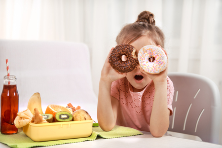 Little beautiful cheerful girl eating a donut and lunch box with fruits at home at the table, the concept of children's foodの写真素材
