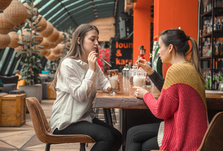 Two happy women are sitting in a cafe, drinking milkshakes, telling each other funny stories, being in a good mood, laughing happily. Best friends have fun together in the cafeの写真素材