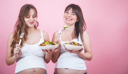 maternity concept, two pregnant women eating fresh salad on a pink colored background, in jeans and a white T-shirt, female friendshipの写真素材