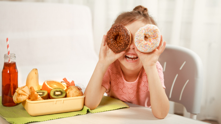Little beautiful cheerful girl eating a donut and lunch box with fruits at home at the table, the concept of children's foodの写真素材