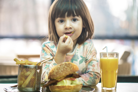 Little cute girl eating a fast food sandwich with fries and orange juice in a cafe. Fast food concept.の写真素材