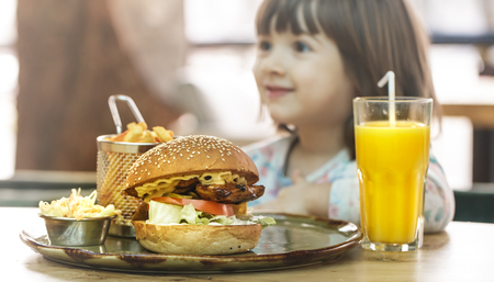 Little cute girl eating a fast food sandwich with fries and orange juice in a cafe. Fast food concept.の写真素材