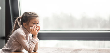 Charming little girl sitting at a wooden table by the window. Upset childの写真素材