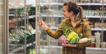Woman in the supermarket. Beautiful young woman holds in hands fresh organic vegetables and opens the fridge in the supermarket. The concept of healthy eating. Harvestの写真素材