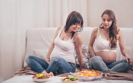 Maternity concept, two pregnant women eating pizza and salad at home on the sofa, friendship and healthy eatingの写真素材