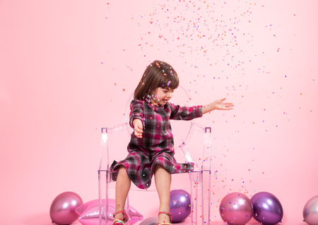 Funny child sitting on a transparent stylish chair on a pink background. Little girl having fun with confetti. The concept of celebration and fun.の写真素材