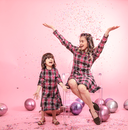 Funny mom and child sitting on a transparent stylish chairs pink background. Little girl and mother having fun with balloons and confetti. The concept of celebration and fun.の写真素材
