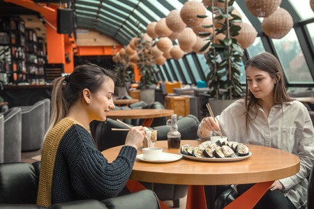 Two happy women sitting in a cafe, eating sushi tell each other funny stories, being in a good mood, laughing happily. Best friends have fun together in the cafeの写真素材