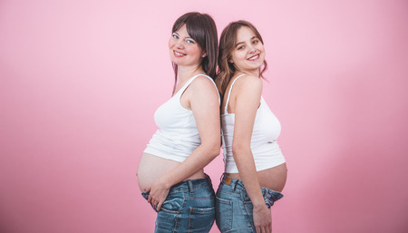 maternity concept, two beautiful pregnant women with an open tummy in jindes and white t-shirts on a colored background, female friendshipの写真素材